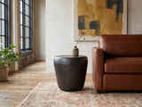 Cervara Rustic Bronze Metal Drum Side Table beside a leather sofa in an industrial loft