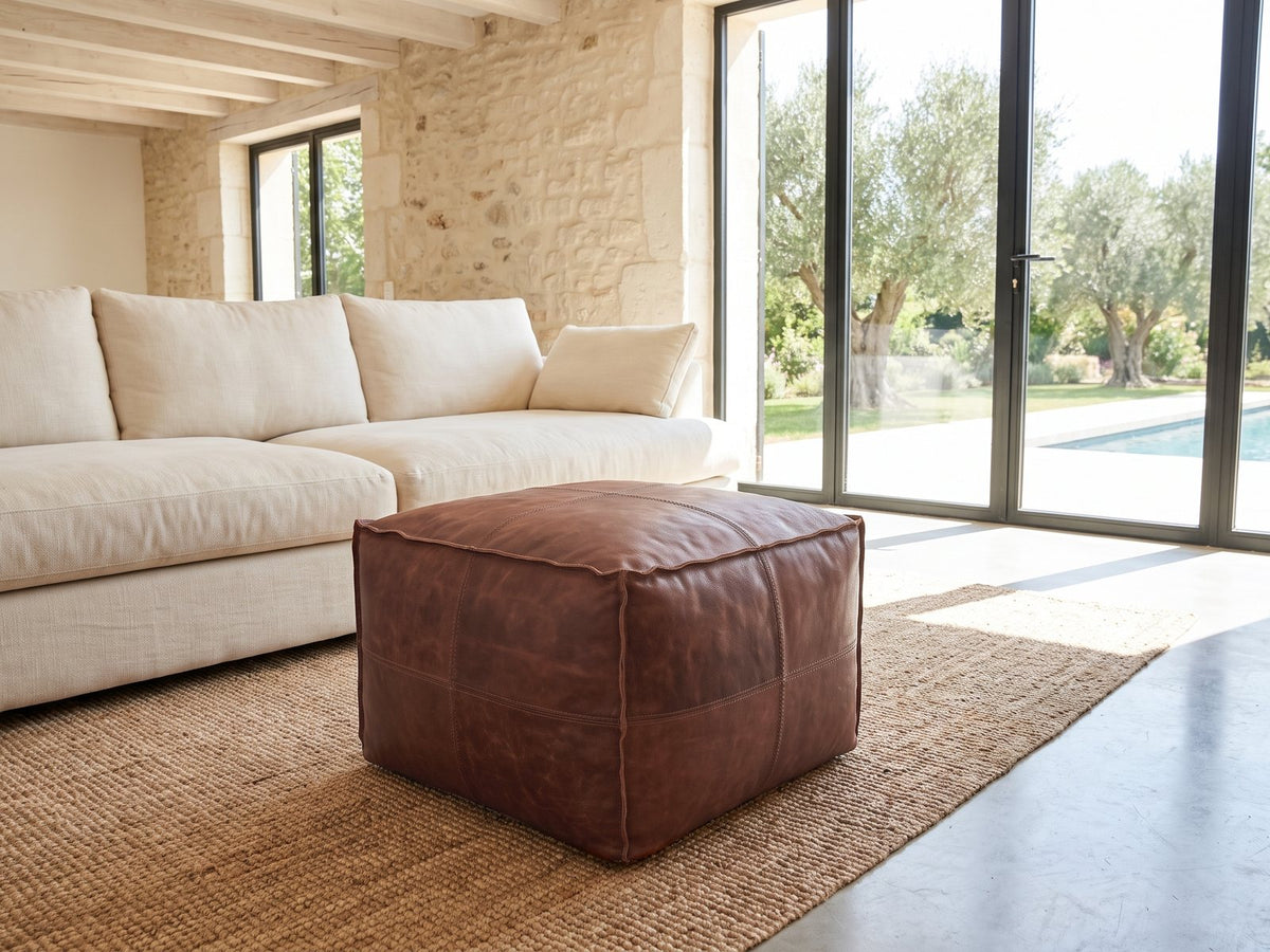Cervo Distressed Brown Genuine Leather Square Pouf on a jute rug in front of a linen sofa in a poolside villa