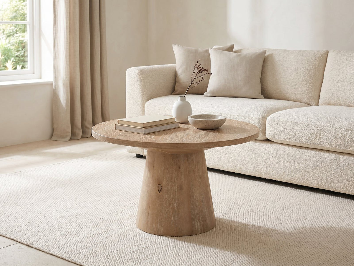 Chiavari Oak Veneer Round Pedestal Coffee Table styled with books, a vase and a stone bowl in front of a cream sofa in a bright plaster room