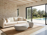 Cilento Grey Fiberglass Pebble Coffee Table and Side Table Set seen from the side, on a jute rug in front of a linen sofa in a poolside villa