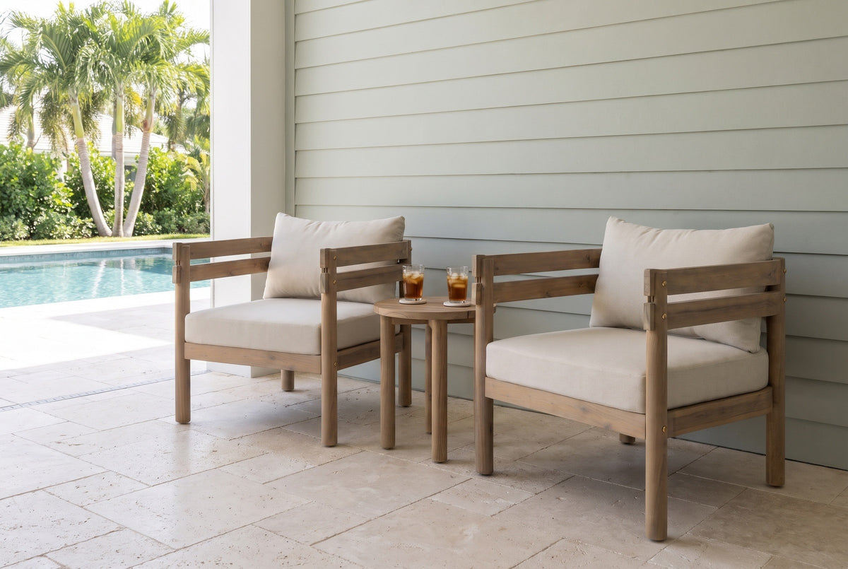 Two Cinisi acacia club chairs on ivory travertine in a covered lanai in Naples, Florida, a pale grey-green shiplap wall and a pool with palms beside (side table and glasses not included)
