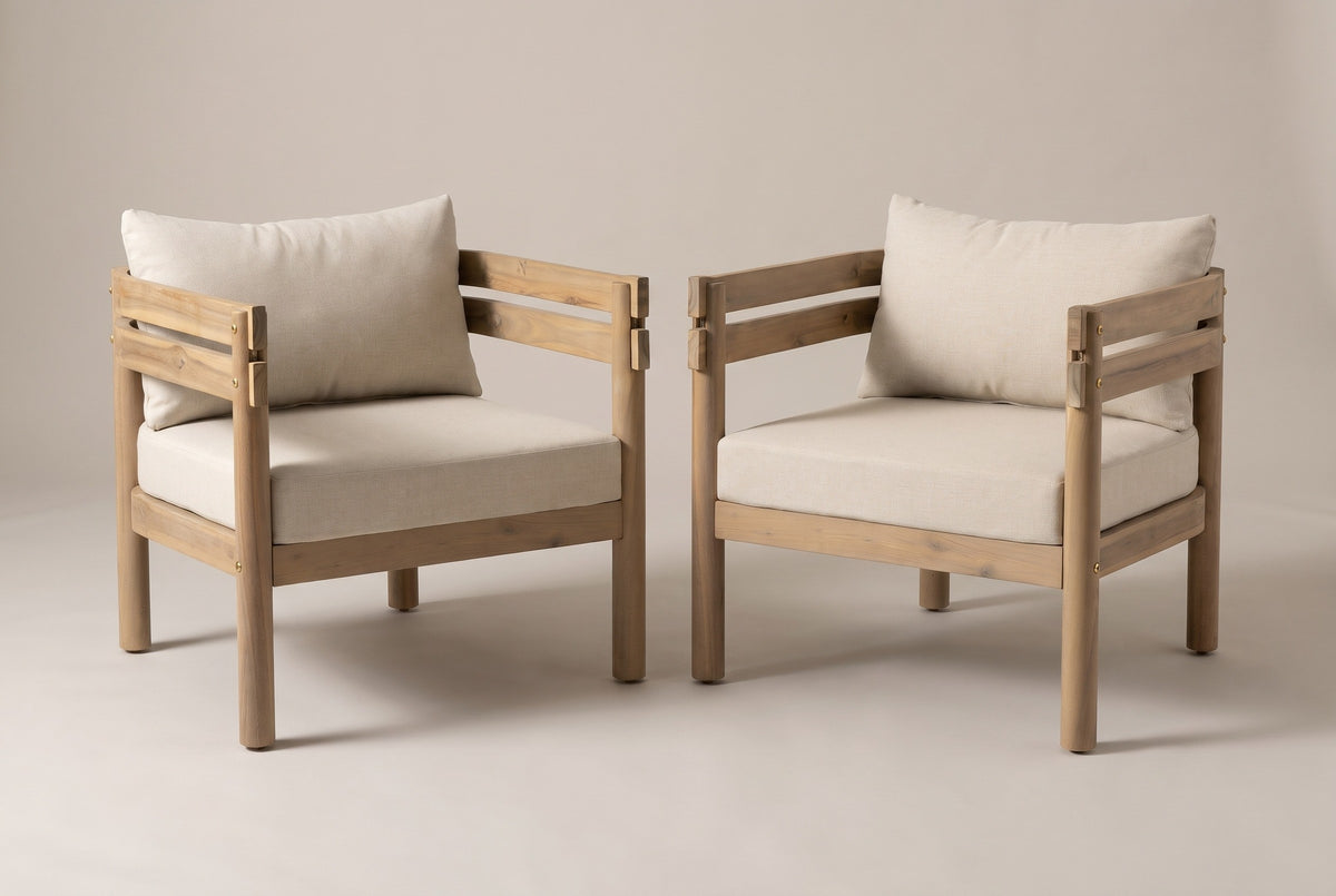 The pair of Cinisi outdoor club chairs in light brown acacia with beige seat and back cushions, three-quarter view on a light studio backdrop
