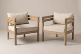 The pair of Cinisi outdoor club chairs in light brown acacia with beige seat and back cushions, three-quarter view on a light studio backdrop