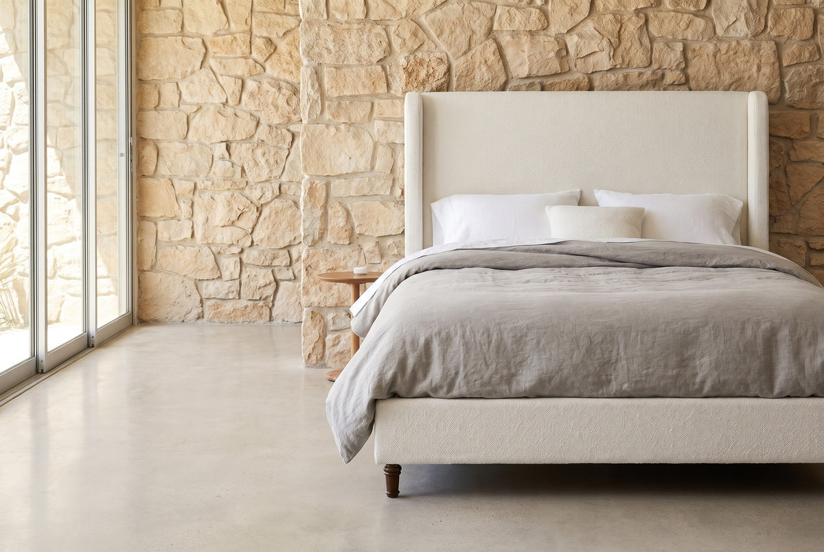 Conversano cream boucle wingback bed against a rough-cut sand-coloured stone wall in a Palm Springs bedroom, a tall glass sliding door at the left