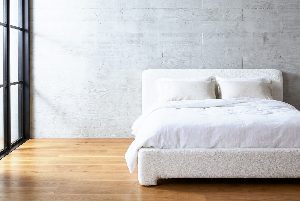 Cupello white boucle platform bed against a pale grey board-formed concrete wall in a Denver bedroom, a tall black steel window at the left