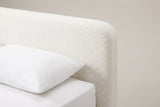 The top corner of the Cupello headboard: a thick white boucle slab with a straight top edge and a softly rounded corner, above a white pillow