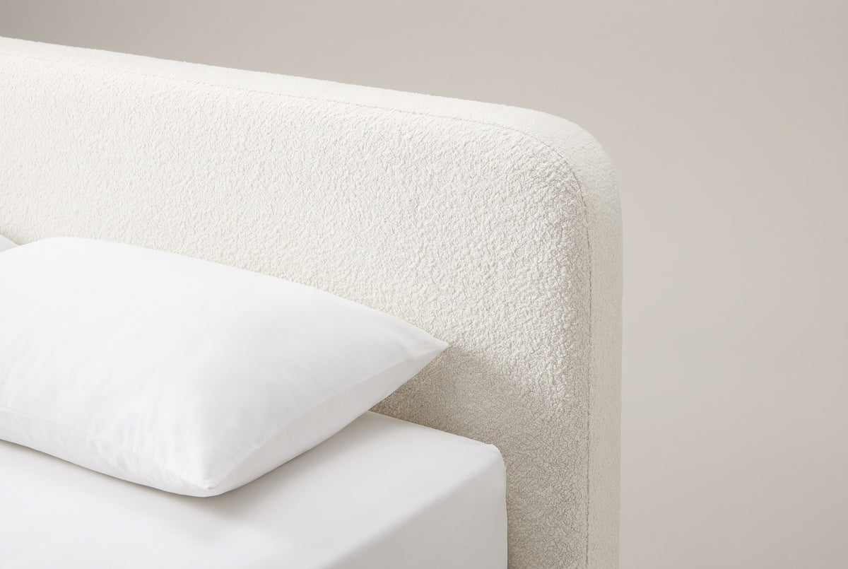 The top corner of the Cupello headboard: a thick white boucle slab with a straight top edge and a softly rounded corner, above a white pillow