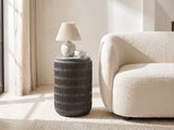 Cutigliano Cast Aluminum Carved Cylinder Side Table with a lamp and a ceramic bowl beside a cream boucle armchair in a bright plaster room