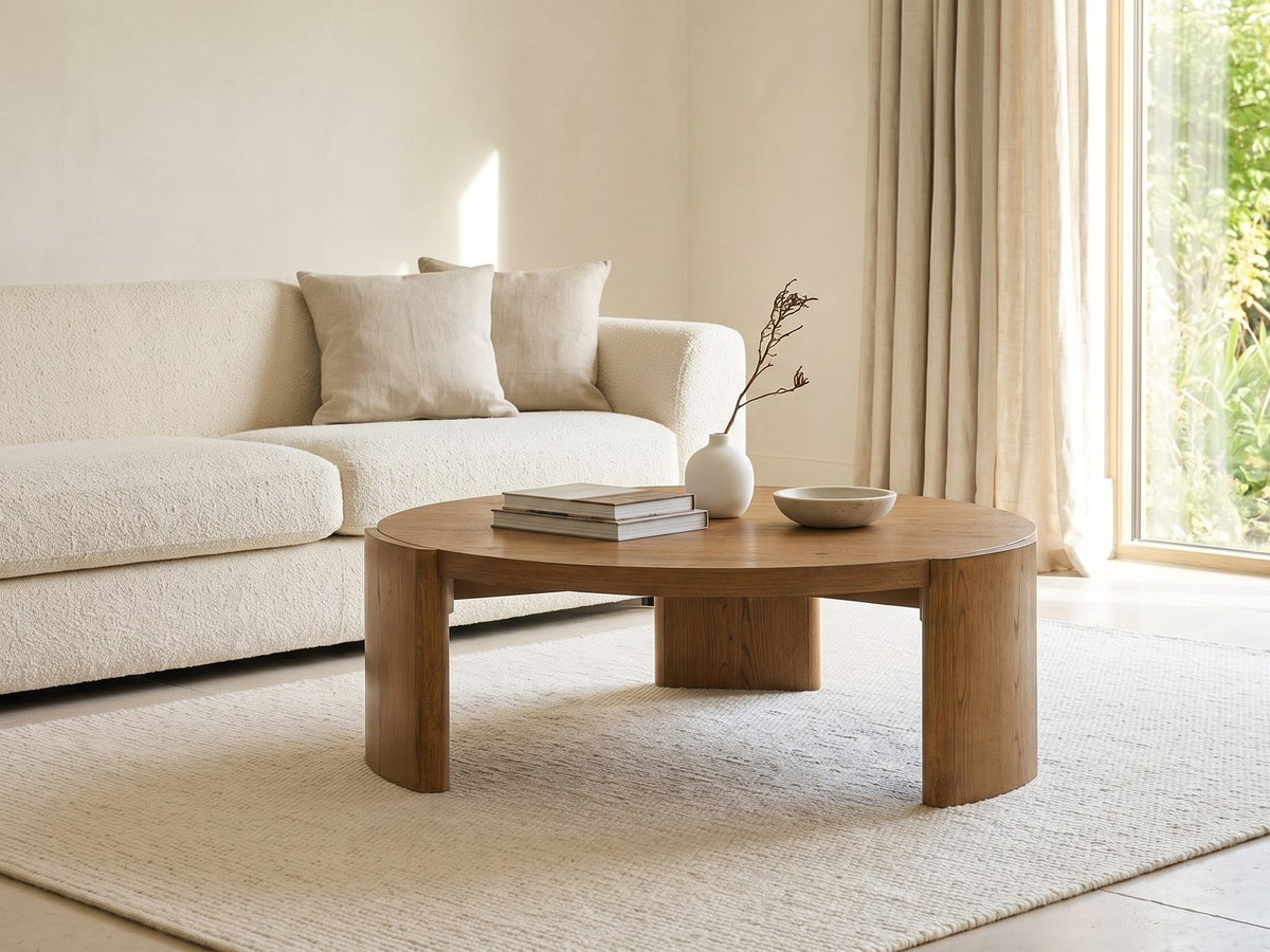 Dogliani 42-Inch Round Walnut-Stained Oak Veneer Coffee Table styled with books, a vase and a stone bowl in front of a cream sofa in a bright plaster room