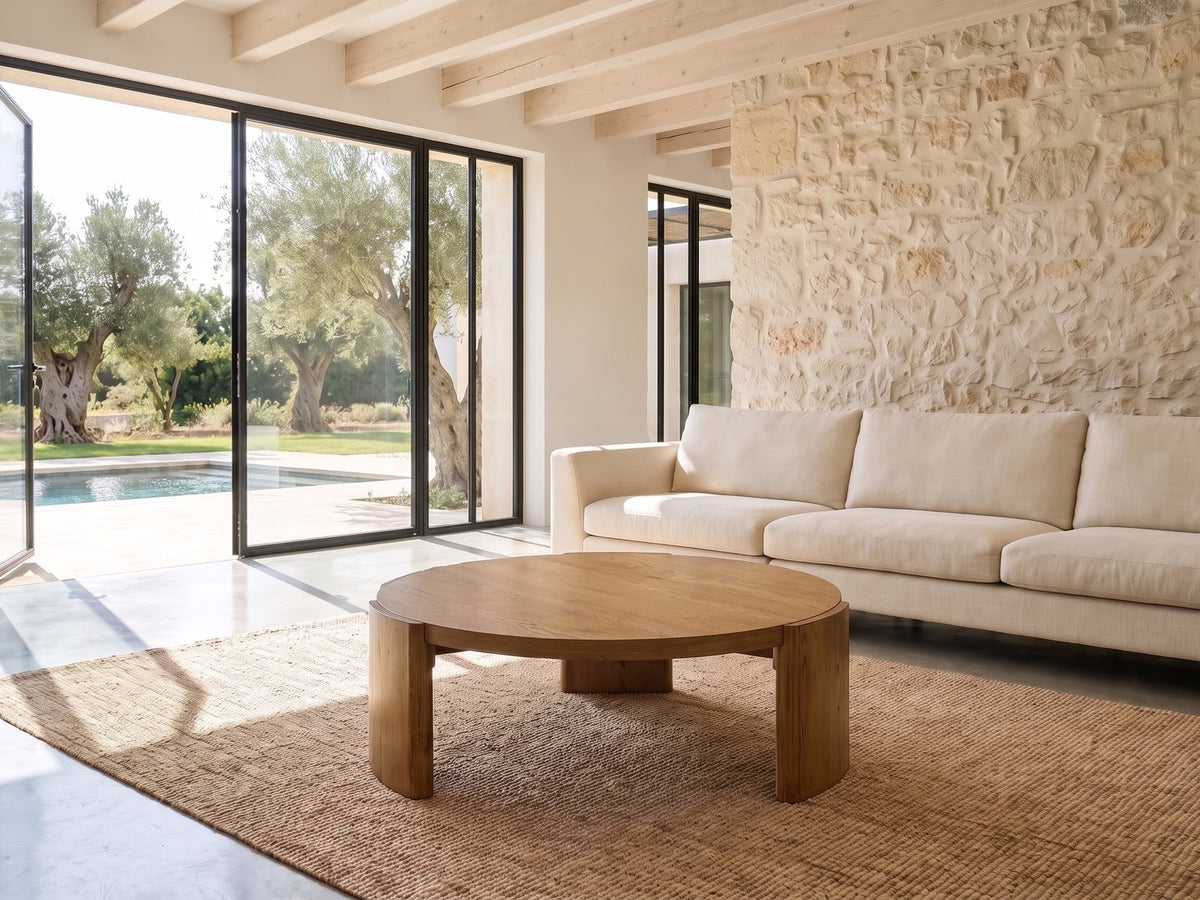 Dogliani 42-Inch Round Walnut-Stained Oak Veneer Coffee Table seen from the side, on a jute rug in front of a linen sofa in a poolside villa