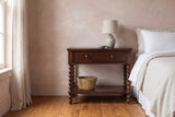 Dorgali Morocco Brown spindle-leg nightstand with a ceramic lamp and a seagrass basket on the shelf beside a bed in a Charleston bedroom with a blush lime-wash plaster wall and a heart-pine floor