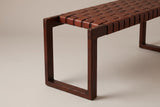 One end of the Fasano bench: the rectangular wood end frame with a small dowel plug in the top rail, and the cognac leather straps wrapping over the front edge beside it