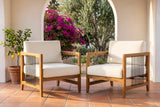Two Ficarra acacia armchairs with ivory cushions on a terracotta tile porch, a whitewashed arch behind with bougainvillea and an olive tree beyond (planter not included)