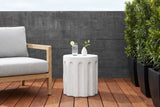 Ficuzza white fluted concrete side table with a glass of sparkling water and a white bud vase beside an oak armchair on an ipe deck in Dallas, Texas, a charcoal board-formed concrete wall behind (chair and decor not included)