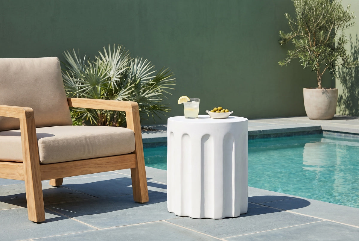 Ficuzza white fluted concrete side table with a glass of lemonade and a dish of olives beside a teak armchair on a bluestone pool terrace in Delray Beach, Florida, a sage-green stucco wall behind (chair and decor not included)