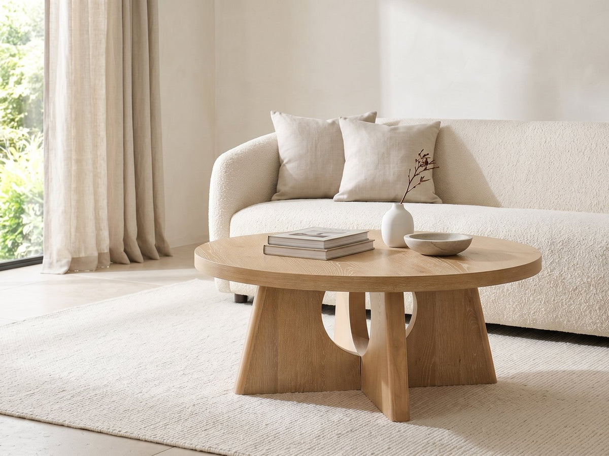 Fiesole Cerused Oak Veneer Round Pedestal Coffee Table styled with books, a vase and a stone bowl in front of a cream sofa in a bright plaster room