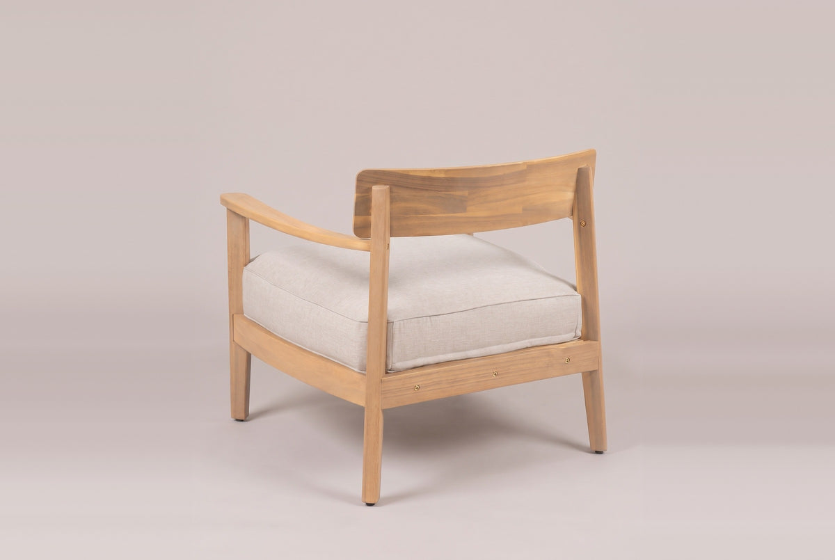 A Furnari club chair from behind: the curved acacia back panel on two square posts, the beige seat cushion and an arm sweeping forward