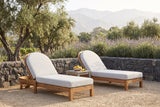 Two Gangi acacia chaise lounges with light grey cushions on a decomposed granite terrace in Ojai, a dark fieldstone wall, lavender and olive trees behind (side table not included)
