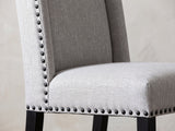 Close-up of the Gerace 72-Inch White Marble V-Trestle Dining Set with 6 Chairs: the winged back and seat of a chair in dove-grey fabric with black nailhead trim