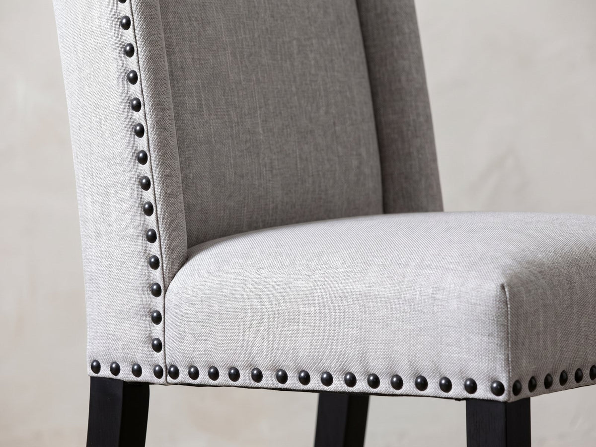 Close-up of the Gerace 72-Inch White Marble V-Trestle Dining Set with 6 Chairs: the winged back and seat of a chair in dove-grey fabric with black nailhead trim