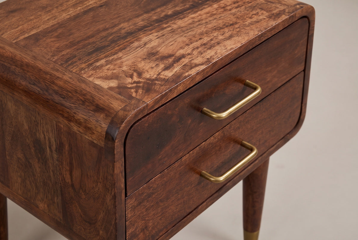 The front corner of the Ghilarza nightstand: the top rolling over a rounded corner of curved mango blocks, two flush drawers with their slim brass U-shaped pulls and the top of a tapered leg