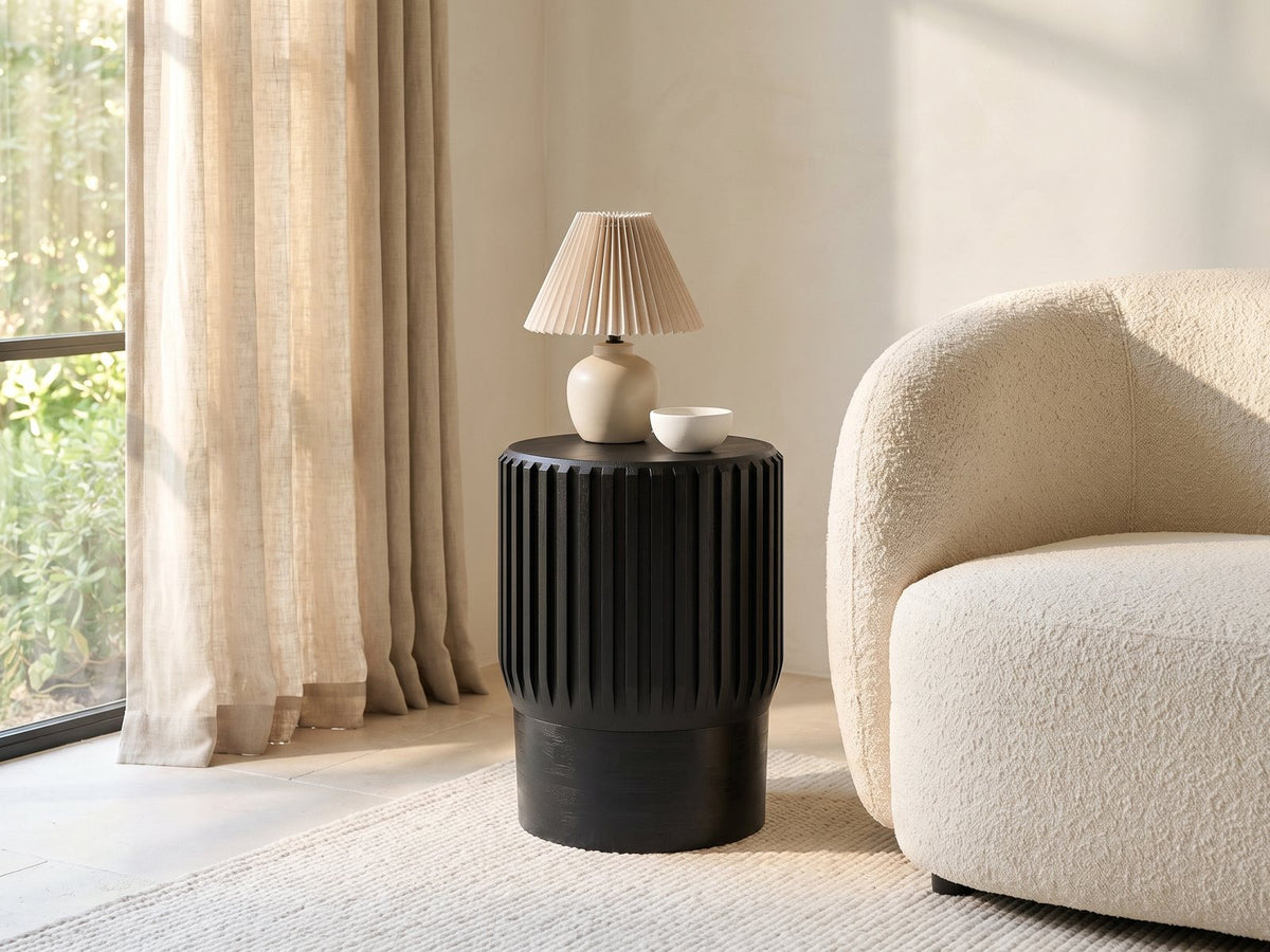Grottole Black Mango Ribbed Drum Side Table with a lamp and a ceramic bowl beside a cream boucle armchair in a bright plaster room