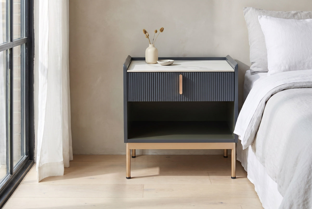 Guspini charcoal fluted nightstand with a white sintered stone top beside a bed in a Chicago loft bedroom with a greige plaster wall and a tall black steel window