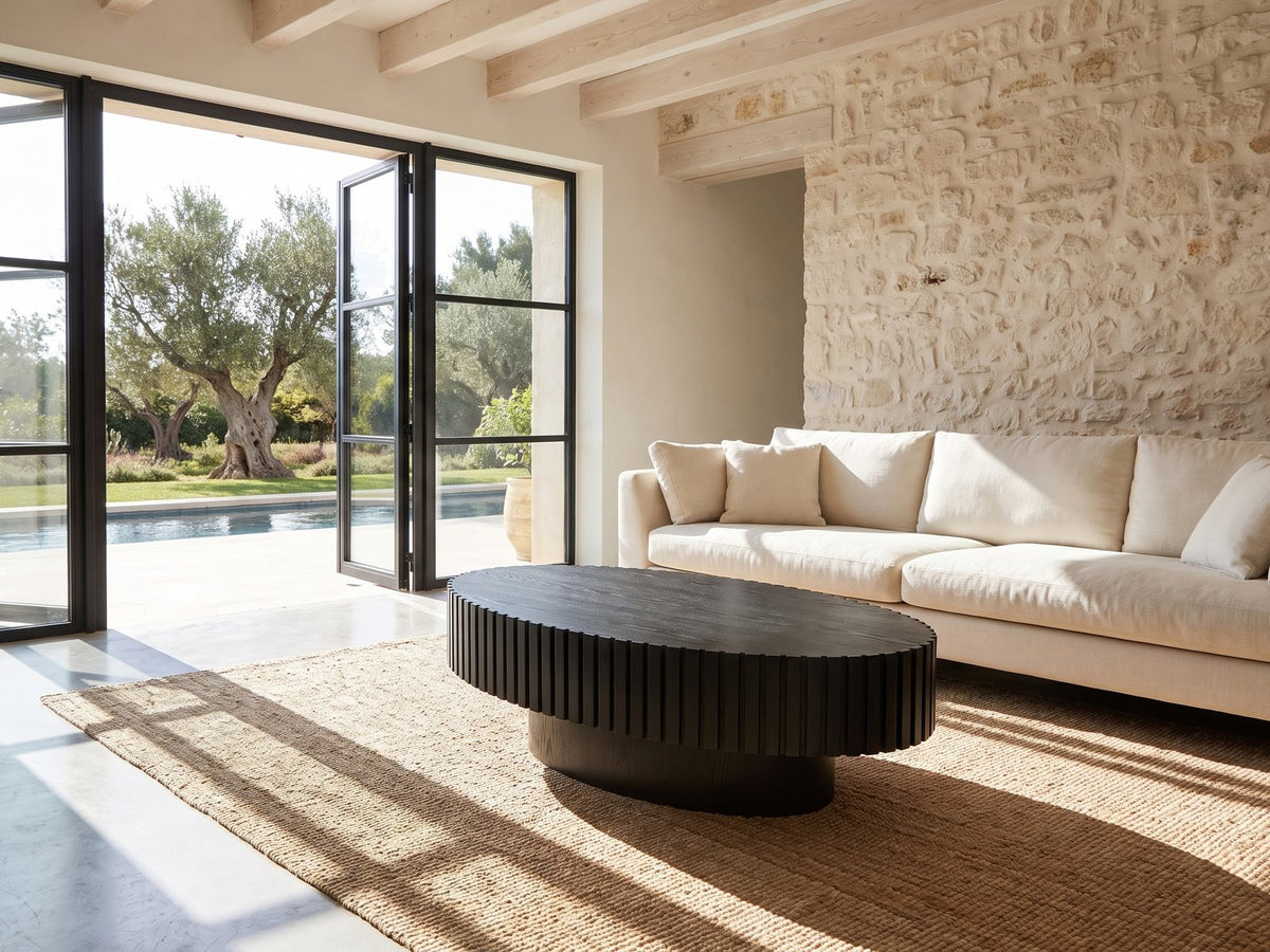Imola Black Ash Oval Pedestal Coffee Table seen from the side, on a jute rug in front of a linen sofa in a poolside villa