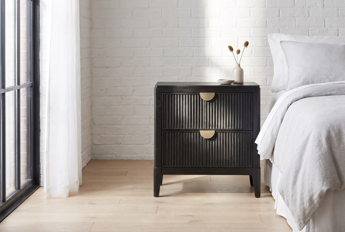 Isili black fluted nightstand beside a bed in a Chicago loft bedroom with a whitewashed brick wall and a tall black steel window