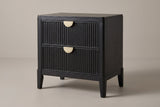 The Isili nightstand in wire-brushed black wood with two fluted drawers, brass half-moon pulls and four square tapered legs, three-quarter view on a light studio backdrop