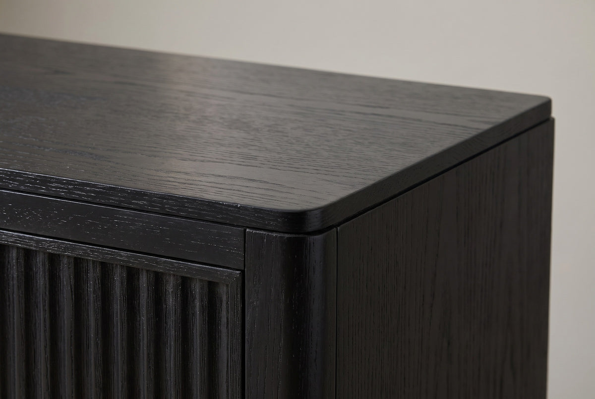 The top corner of the Isili nightstand: a thin black wood top with a rounded front corner, the rounded corner post and the top of a fluted drawer front