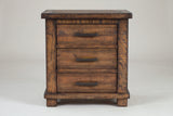 The Ittiri three-drawer nightstand in reclaimed pine, front view on a light studio backdrop: a framed plank top, three drawers with bronze bar pulls, wide corner posts, a base rail and block feet