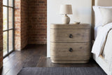 Lanusei knotty oak nightstand with a ceramic lamp beside an upholstered bed in a Chicago loft bedroom with exposed brick, a steel-framed window and a dark wood floor