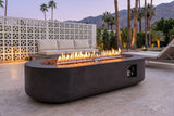 Licata oval concrete fire pit table lit at blue hour on travertine pavers in a Palm Springs patio, a white breeze-block wall, palms and mountains behind (sofa and lounge chair not included)