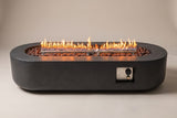 The Licata fire pit table seen from the long side: the low concrete body with rounded ends, the lit burner over volcanic rock and the control niche near the right-hand end