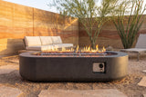 Licata oval concrete fire pit table lit at golden hour on decomposed granite in a Tucson desert patio, a layered rammed-earth wall, a palo verde tree and an ocotillo behind (sofa and lounge chair not included)