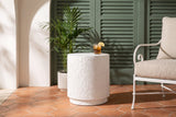 Linguaglossa white concrete drum side table on terracotta hexagon tiles in a Palm Beach loggia, sage green shutters and a palm behind, an iced tea on top, a white iron armchair beside it (chair and decor not included)
