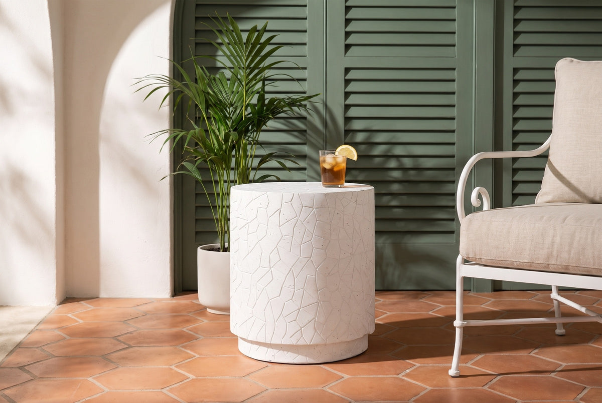 Linguaglossa white concrete drum side table on terracotta hexagon tiles in a Palm Beach loggia, sage green shutters and a palm behind, an iced tea on top, a white iron armchair beside it (chair and decor not included)