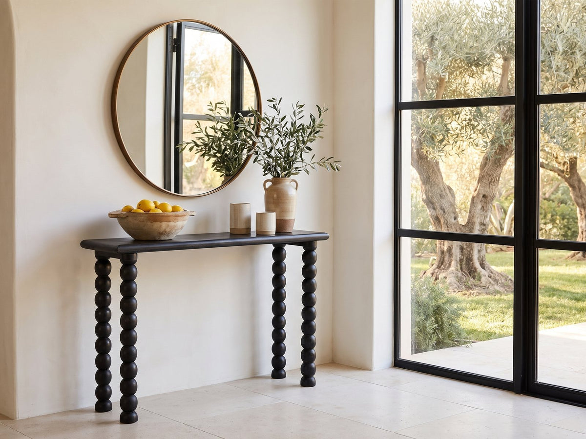 Lizzano Black Mango Wood Bobbin Console Table under a round bronze mirror beside a glass wall onto an olive garden in a sunlit villa