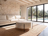 Lucignano Whitewashed Mango Drum Coffee Table seen from the side, on a jute rug in front of a linen sofa in a poolside villa
