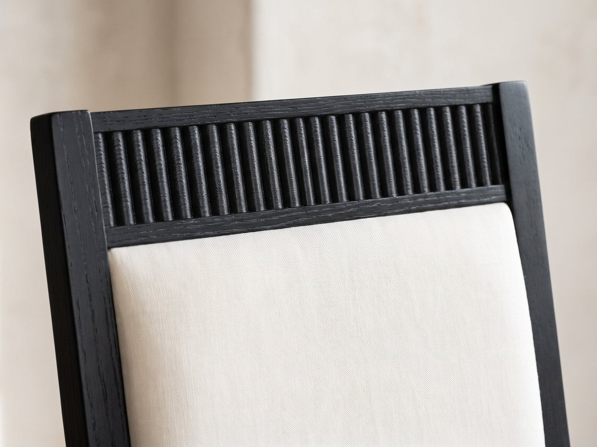 Close-up of the Lungro 82-Inch Reeded Black Oak Extension Dining Set with 4 Chairs: the reeded black crest rail and ivory upholstered back of a chair