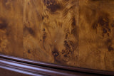 Close-up of the poplar burl veneer on a Luras drawer front: golden amber and chestnut figure with dark swirling eyes and thin seams between the leaves, above the dark cherry moulding