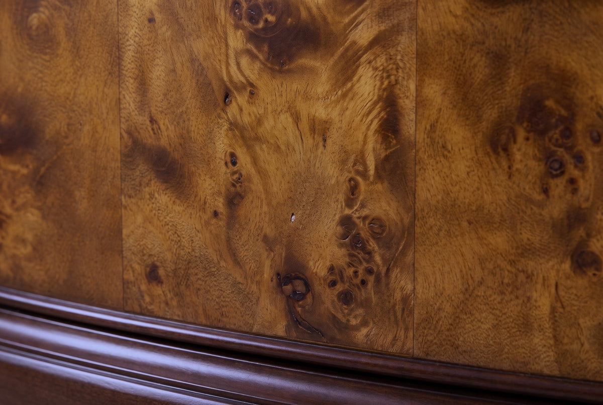 Close-up of the poplar burl veneer on a Luras drawer front: golden amber and chestnut figure with dark swirling eyes and thin seams between the leaves, above the dark cherry moulding
