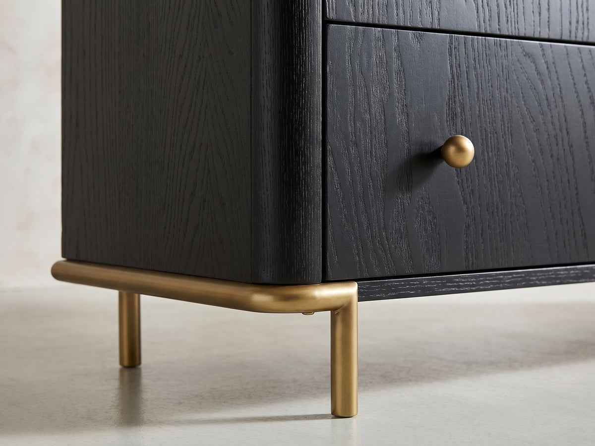 Close-up of the Malcesine 63-Inch Wire-Brushed Black 8-Drawer Dresser: the soft-gold metal sled under one end, the rod curving round the corner into a slim round leg