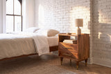 Mamoiada golden-brown mango nightstand against a whitewashed brick wall in a Brooklyn bedroom with a herringbone floor and a tall window