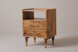 The Mamoiada open-shelf nightstand in golden-brown mango with a low rim on the top, an open cubby, two drawers with slim brass bar pulls and four tapered legs with brass caps, three-quarter view on a light studio backdrop