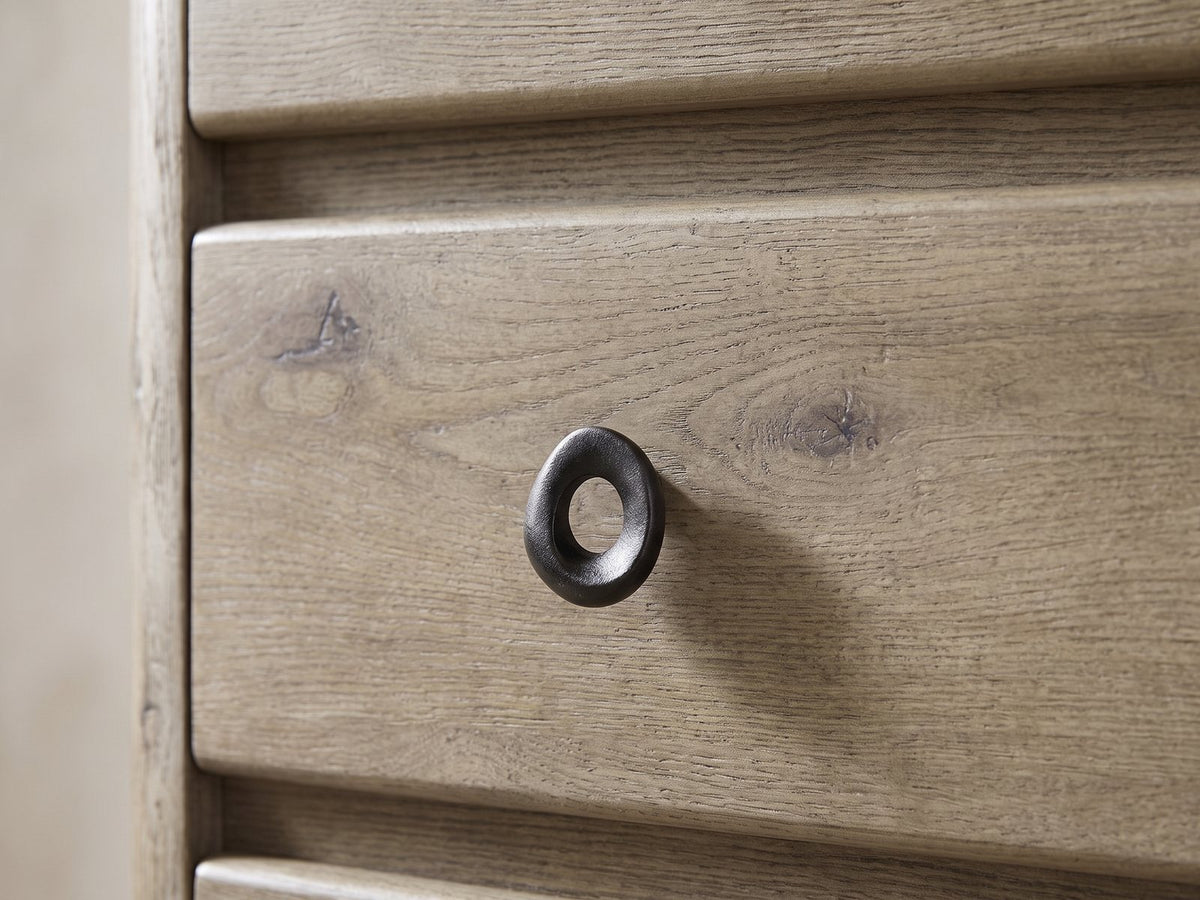 Close-up of the Maniago 42-Inch Weathered Natural Oak 5-Drawer Chest: a dark cast-iron oval ring pull on a knotty oak drawer front beside the rounded case edge