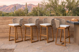 Four Mascali acacia bar stools with curved wicker backs and pale cushions in a row on a flagstone terrace in Tucson, a rammed-earth wall and outdoor kitchen behind and saguaros and the Catalina mountains beyond (outdoor kitchen not included)