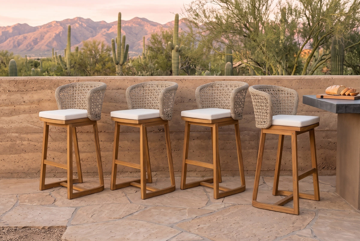 Four Mascali acacia bar stools with curved wicker backs and pale cushions in a row on a flagstone terrace in Tucson, a rammed-earth wall and outdoor kitchen behind and saguaros and the Catalina mountains beyond (outdoor kitchen not included)
