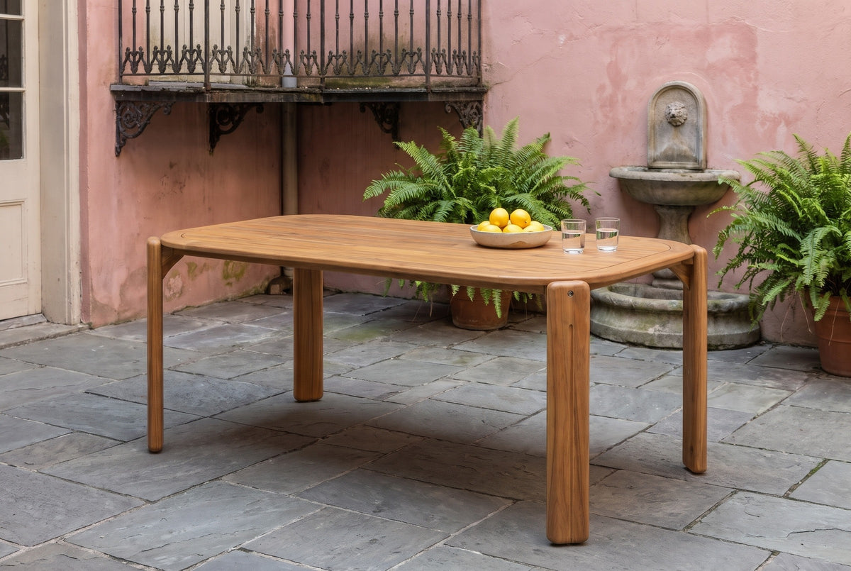 Mazara acacia dining table in a New Orleans French Quarter courtyard with a rose-pink plaster wall, a wrought-iron balcony, ferns and a stone fountain (chairs not included)
